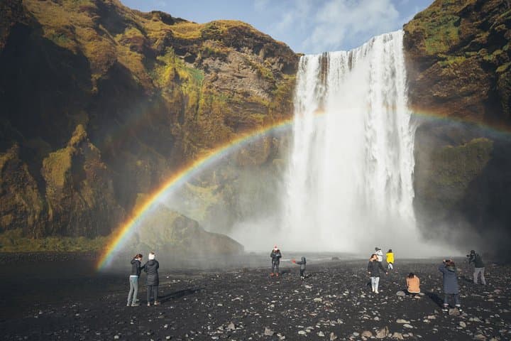 Rainbow across Skogafoss waterfall, seen from a black-sand beach.