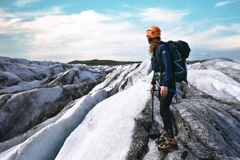 A person in a blue jacket, wearing crampons and carrying an ice axe, looks at the frozen landscape around her during a glacier hike at Solheimajokull Glacier.