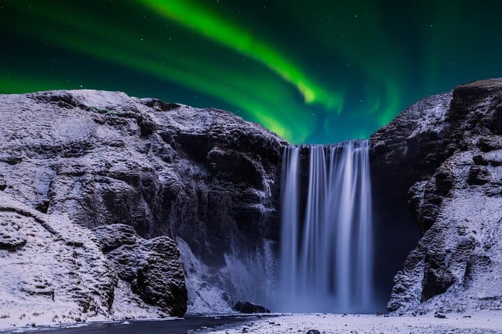 Skogafoss Waterfall flows beneath stunning northern lights in the night sky.